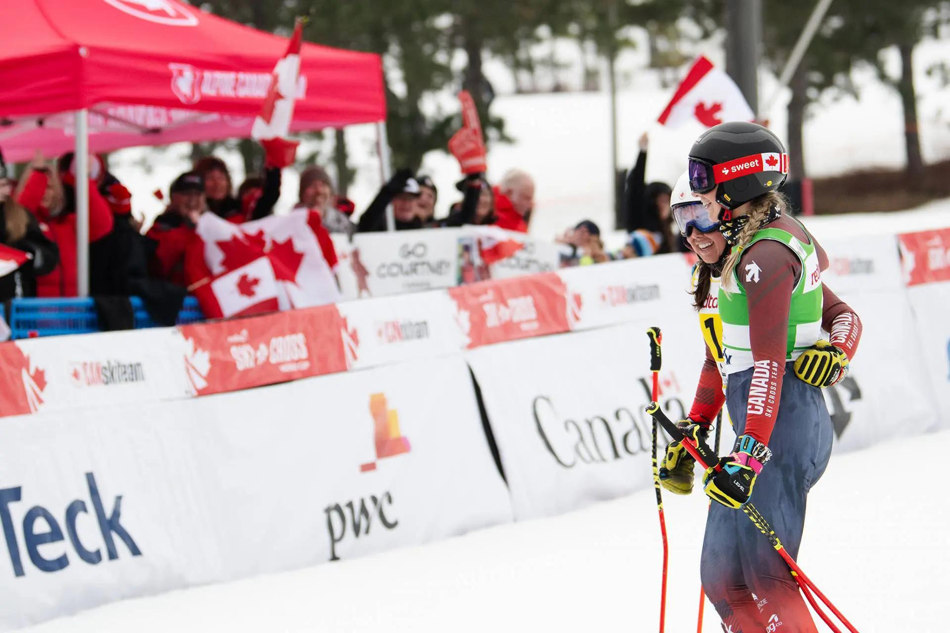 deux skieuses ensemble devant des supporters agitant des drapeaux canadiens